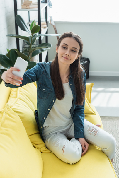 attractive smiling girl sitting on sofa and taking selfie with smartphone - Zdjęcie, obraz