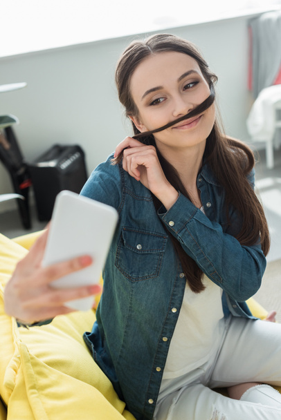 adolescente sonriente haciendo bigote falso de pelo mientras toma selfie con teléfono inteligente en casa
 - Foto, imagen