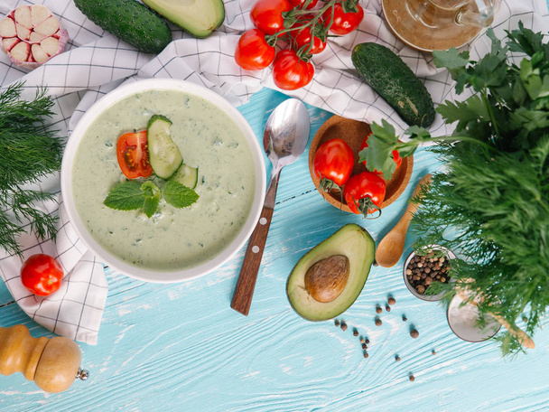 Kom met de crème van avocado soep met tomaten en komkommers op blauwe houten achtergrond - Foto, afbeelding