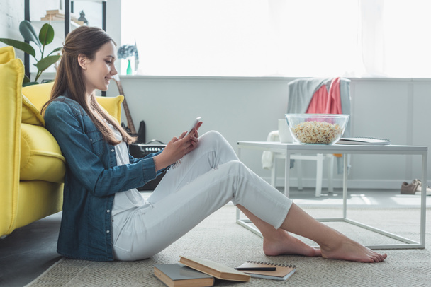 side view of smiling teenage girl using smartphone while sitting on carpet with books at home - Photo, Image
