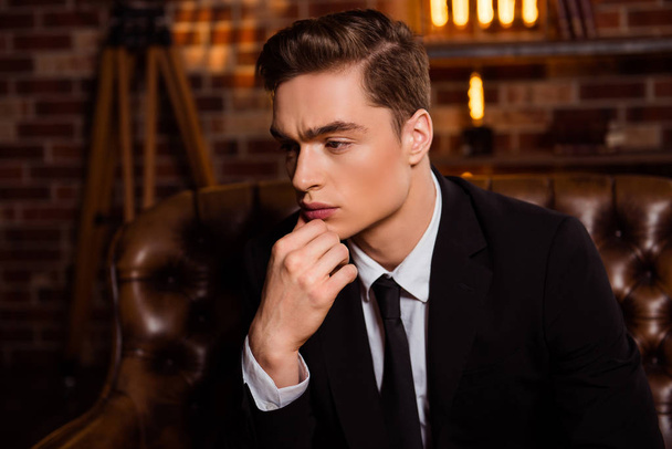 Close up portrait, half-turned face of harsh, brutal, perfect man in black suit with tie sitting on leather couch, holding hand on chin, with thoughtful expression looking to the side - Zdjęcie, obraz