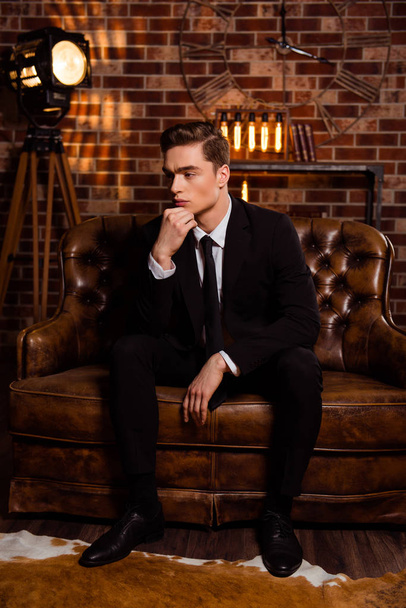 Manly, virile man in black suit with tie sitting on leather sofa over brick wall, holding hand on chin, with thoughtful expression looking to the side - Foto, afbeelding