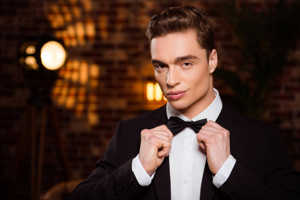 Portrait of manly, harsh, perfect man in black suit, preparing for date, holding hands on bowtie over brown blurred brick wall, looking at camera - Foto, Imagem