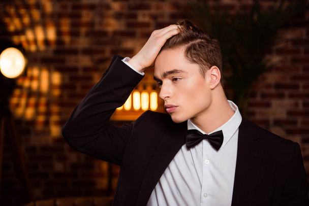 Half turned face portrait of cool macho, confident man in black suit with bowtie touching his perfect hair over brown brick wall, looking to the side - Fotó, kép