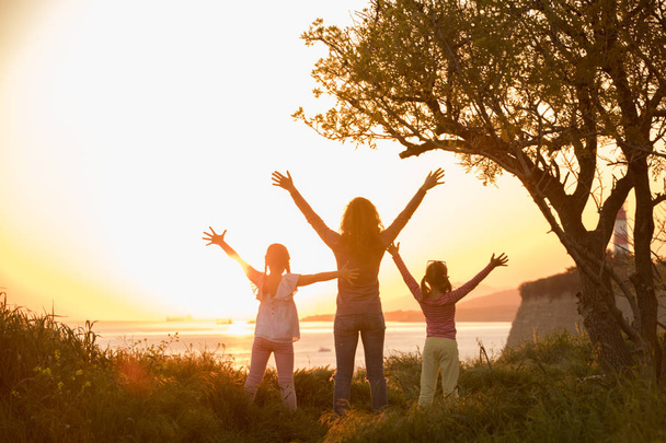 Silhueta de mãe com duas filhas pequenas olhando para o pôr do sol e água
 - Foto, Imagem