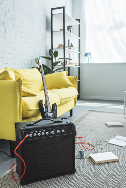 Elektrická kytara a hlasitý reproduktor s smartphone stojící na podlaze s knihami poblíž pohovka - Fotografie, Obrázek