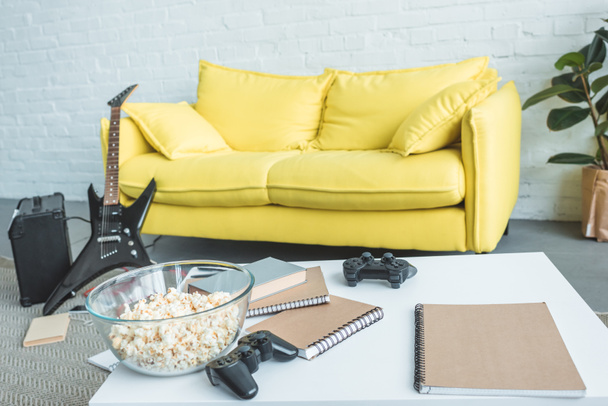 popcorn, joysticks and copybooks on table, electric guitar near couch behind - Photo, Image