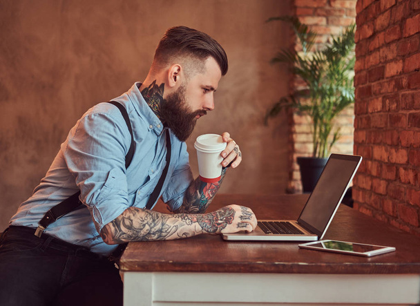 Handsome tattooed hipster in a shirt and suspenders sitting at the desk, working on a laptop, holds takeaway coffee in an office with loft interior. - Фото, изображение