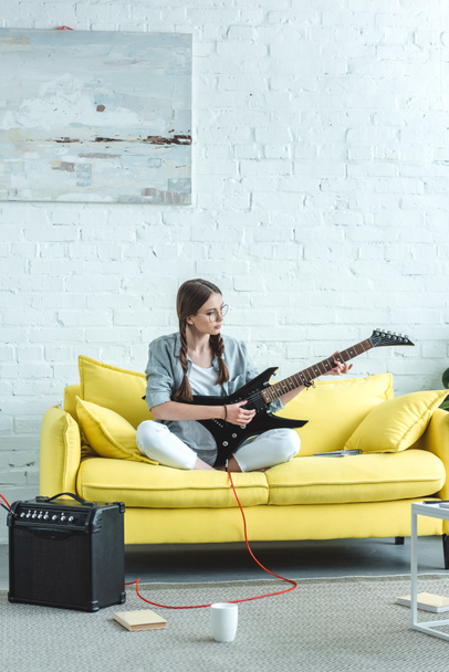 beautiful teen girl playing electric guitar on sofa in living room - Foto, afbeelding