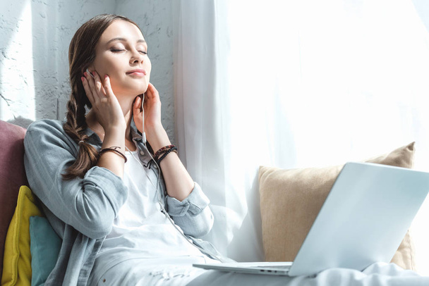 kaunis teini tyttö käyttää kannettavaa tietokonetta ja kuuntelee musiikkia kuulokkeet ikkunalaudalla
 - Valokuva, kuva