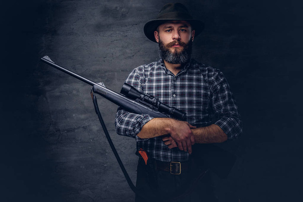 Handsome bearded hunter traveler in a fleece shirt and hat holds rifle with a sight. Isolated on a dark background. - 写真・画像