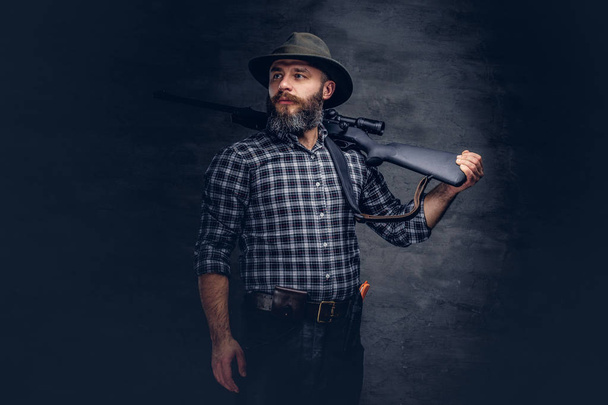 Бородатий мисливець у флісовій сорочці та капелюсі тримає на плечі гвинтівку з прицілом, дивлячись геть. Ізольовані на темному тлі
. - Фото, зображення