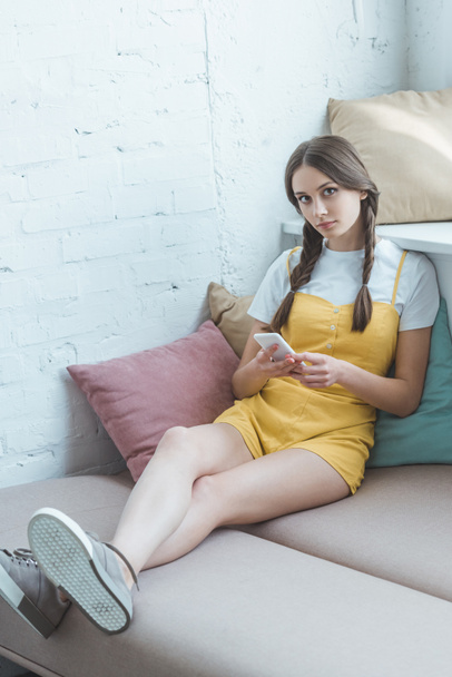 schöne Teenie-Mädchen mit Zöpfen mit Smartphone und sitzt auf dem Sofa - Foto, Bild