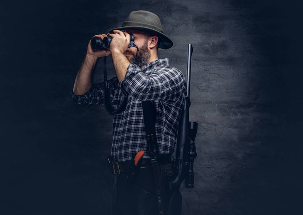 Portrait of a bearded hunter traveler tries to find prey while looking through binoculars. Isolated on a dark background. - Fotografie, Obrázek