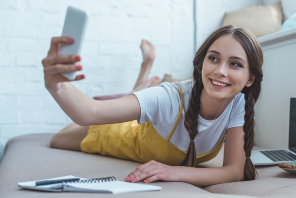 smiling girl taking selfie on smartphone while lying on sofa with copybook and laptop  - Фото, изображение