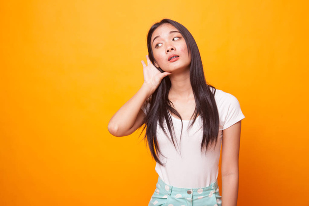 Beautiful young Asian woman listen to something on bright yellow background - Photo, Image