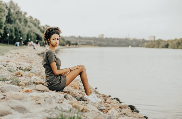 Charming African-American girl is sitting on a stony quay of a river with the park and a walkway behind her; young curly Brazilian female is sitting on a stone embankment on a summer evening - Foto, Imagem