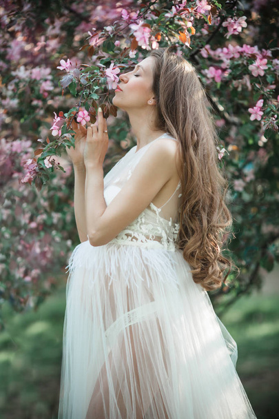 Pregnant pretty woman is wearing white fashion dress posing in pink blossom apple garden, summer time - Photo, Image