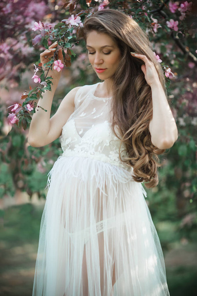 Pregnant pretty woman is wearing white fashion dress posing in pink blossom apple garden, summer time - Photo, Image