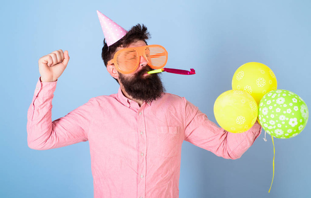 Cheerful bearded man in birthday cap blowing party whistle, fun time, happiness concept. Artist in big crazy glasses holding colorful balloons. Man with hipster beard and mustache celebrating - Photo, Image