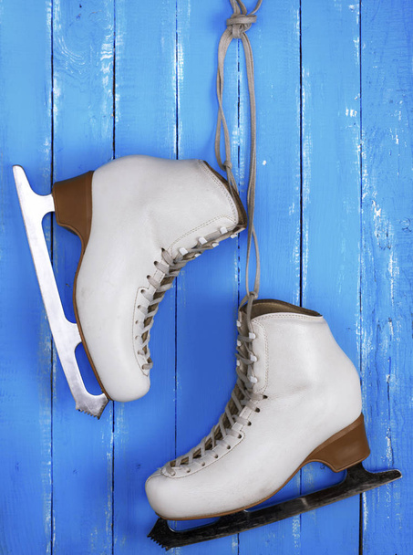 par de patines femeninos de cuero blanco para patinaje artístico colgando de un fondo de madera azul
 - Foto, imagen