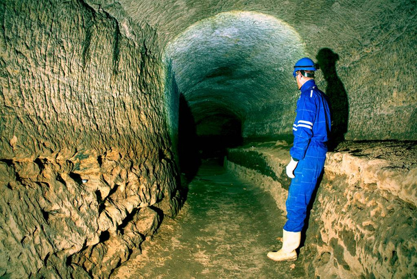 Trabajo subterráneo. Obrero encorvado en un azul en general y un casco de seguridad se encuentra en el túnel subterráneo. El personal revisa la mina subterránea sin apoyo
. - Foto, Imagen