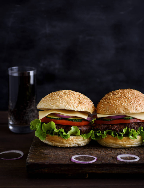 two burger with meat, vegetables and cheese, round biscuit made of wheat flour with sesame, behind the glass of a drink - Foto, afbeelding