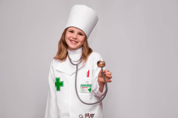 Hymyilevä pikkutyttö lääketieteellisessä univormussa pitäen stetoskooppia ja katsoen kameraa, joka on eristetty valkoisella
 - Valokuva, kuva