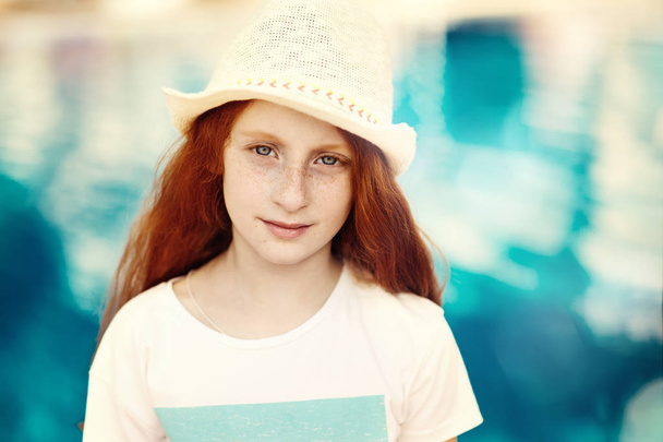 Close-up facial portrait of beautiful little child girl with red hair and freckles standing in front of the pool - 写真・画像