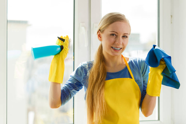 Young woman in yellow gloves cleaning window with blue rag and spray detergent. Spring cleanup, housework concept - Fotoğraf, Görsel