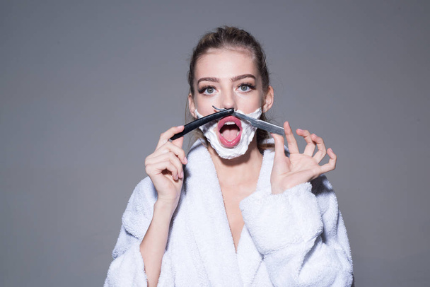 Lady play with sharp blade of straight razor. Barber and shaving concept. Woman with face covered with foam holds straight razor in hand. Girl on smiling face wears bathrobe, grey background - Фото, изображение