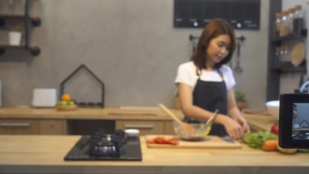 Jovem mulher asiática na cozinha gravação de vídeo na câmera. Sorrindo asiático mulher trabalhando em comida blogger conceito com frutas e legumes na cozinha
. - Filmagem, Vídeo