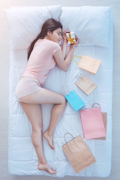 Joyful brunette holding gift box while sleeping in bed - Zdjęcie, obraz