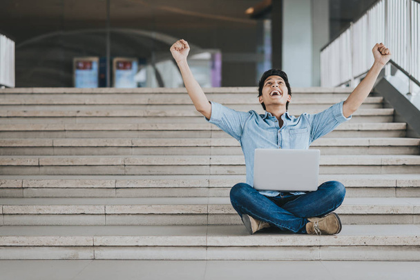 Happy opgewonden Aziatische man holding laptop en het verhogen van zijn arm omhoog te vieren van succes of prestatie. - Foto, afbeelding