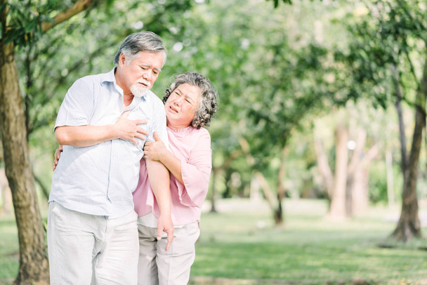 Senior asiatico uomo holding suo petto e sentimento dolore suffering da cuore attacco all'aperto a il parco mentre suo moglie sentimento preoccupazione
 - Foto, immagini