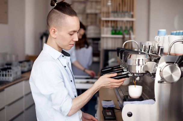 A young men Barista with a stylish hairstyle works in a cafe and brews coffee in the coffee machine holding the handle of the filter holder. Coffee flows into the Cup - Zdjęcie, obraz