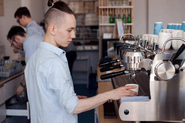 A young guy works as a Barista, he stands at the coffee machine and fills a Cup with hot coffee - 写真・画像