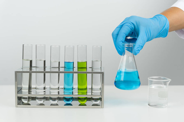 лабораторное место, ученый, держащий синюю жидкую колбу, пробирки в стойке и стакан на столе
 - Фото, изображение