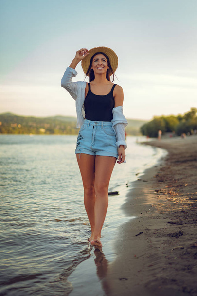Joven mujer sonriente en sombrero de paja caminando al atardecer en la orilla del río
 - Foto, imagen