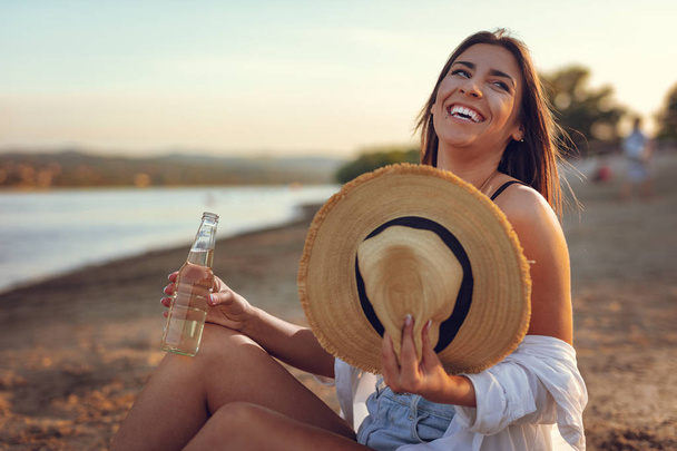 Mladá žena s slamák pít pivo na pláži při západu slunce - Fotografie, Obrázek