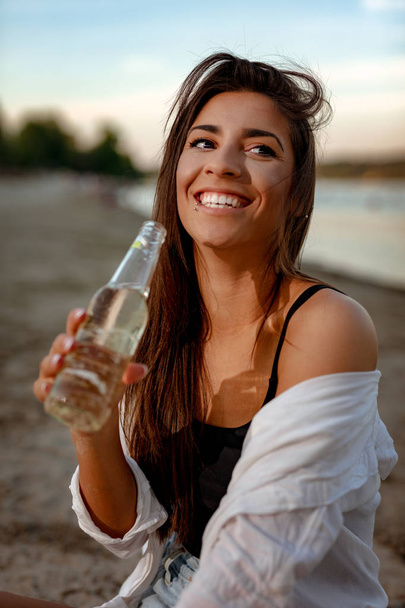 夕暮れ時のビーチでビールを飲むわらの帽子を持つ若い女性 - 写真・画像