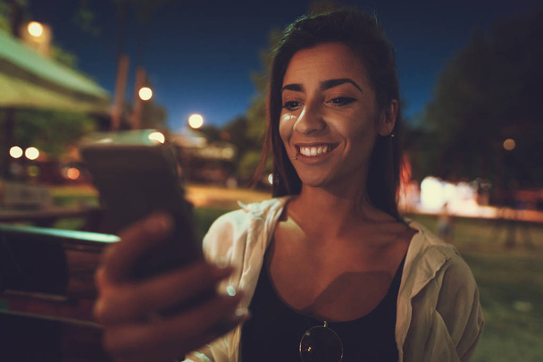 Jonge lachende vrouw surfen netto met smartphone op moment van de avond op de rivier bank - Foto, afbeelding