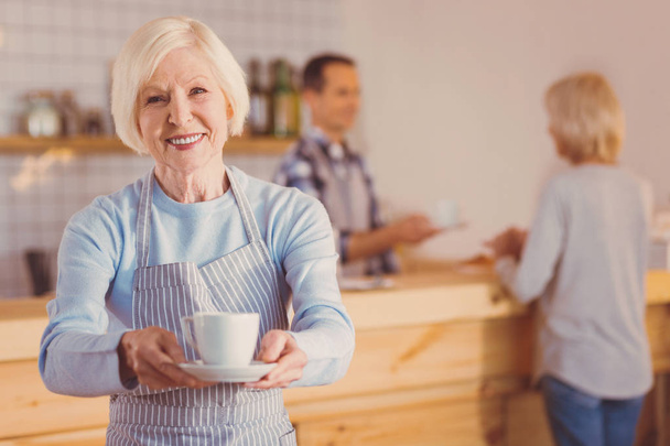 Charming elderly barista posing with a cup of coffee - Fotoğraf, Görsel