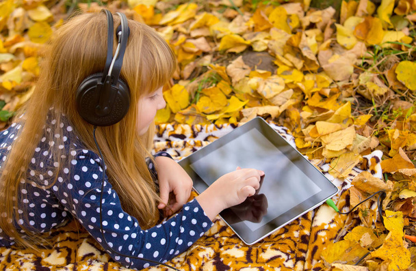 beautiful little girl with earphones using tablet pc in autumn park - Фото, изображение