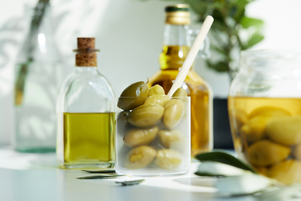 verre avec cuillère et olives vertes, bocal, diverses bouteilles d'huile d'olive aromatique et branches sur table blanche
 - Photo, image