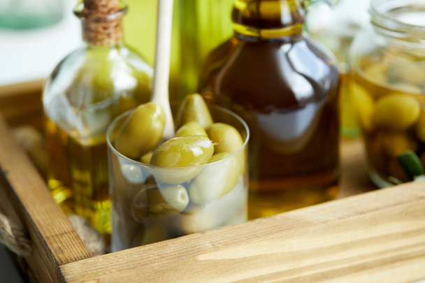 close up view of glass with spoon and green olives, jar, various bottles of aromatic olive oil on wooden tray - Photo, Image