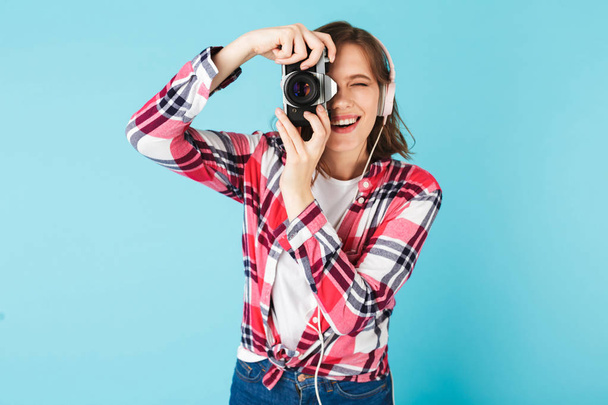 Portrét mladé dámy ve sluchátkách prohlédl v malé retro fotoaparát při pořizování fotografie růžovém pozadí - Fotografie, Obrázek