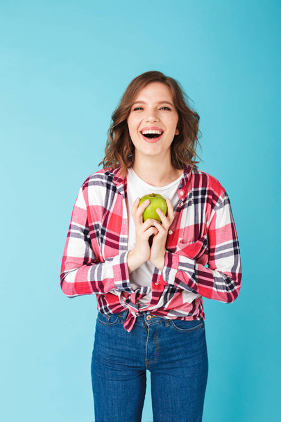 Portrait of beautiful smiling lady standing in shirt and jeans with green apple in hands and joyfully looking in camera on over pink background  - Photo, image