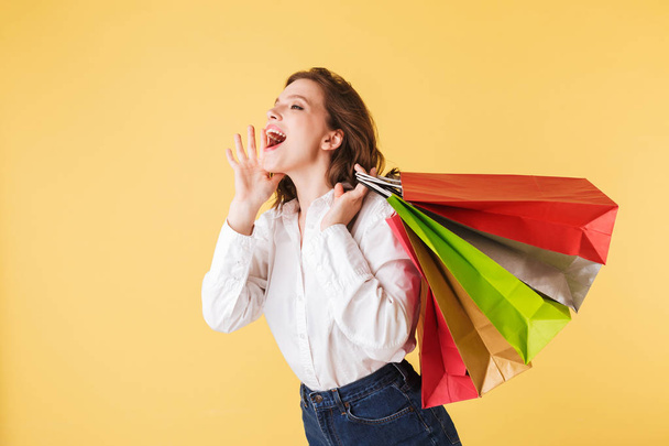 Portrait of young lady in shirt and denim shorts standing with colorful shopping bags in hands and screaming while happily looking aside on over pink background - Foto, Imagem