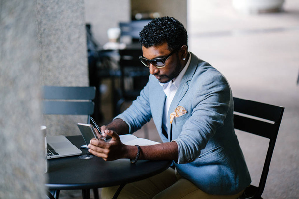 An Indian Asian man is sitting at a coffee shop with his phone and  notebook.  He is dressed in a casual suit. - Photo, Image